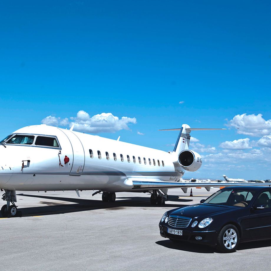 A private jet is parked on an airport tarmac under a blue sky with scattered clouds. A black Mercedes-Benz sedan is parked in front of the jet.